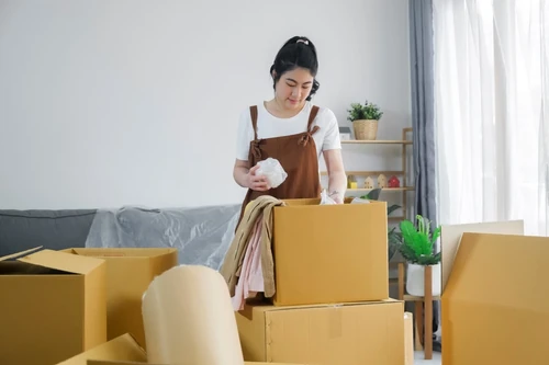 A woman packing clothes and household items into cardboard boxes while preparing for a move in a living room.